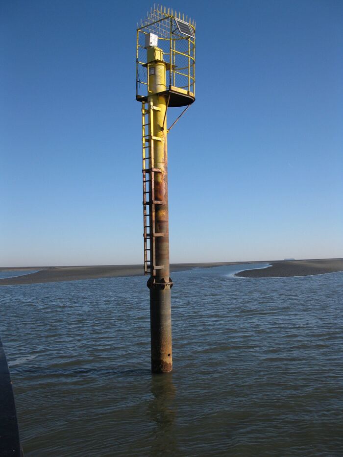 Begehbarer Hilfspegel-Turm im Wattenmeer: massive gelbe Pegelstation mit Antennen­käfig, ausgelegt für hohe Tidenhübe und Sturmlasten.