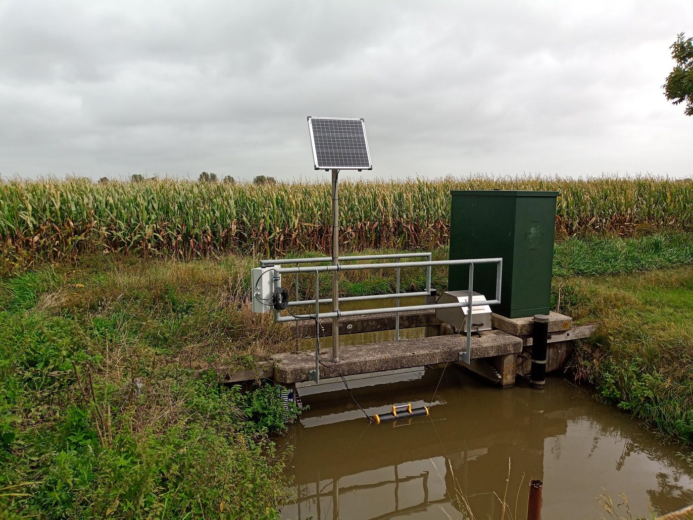 Nitratmätstation vid ett dike: apparatskåp med Aquatos Web LTX, solpanel på en mast och sonden i den gul-svarta flytkroppen i vattnet